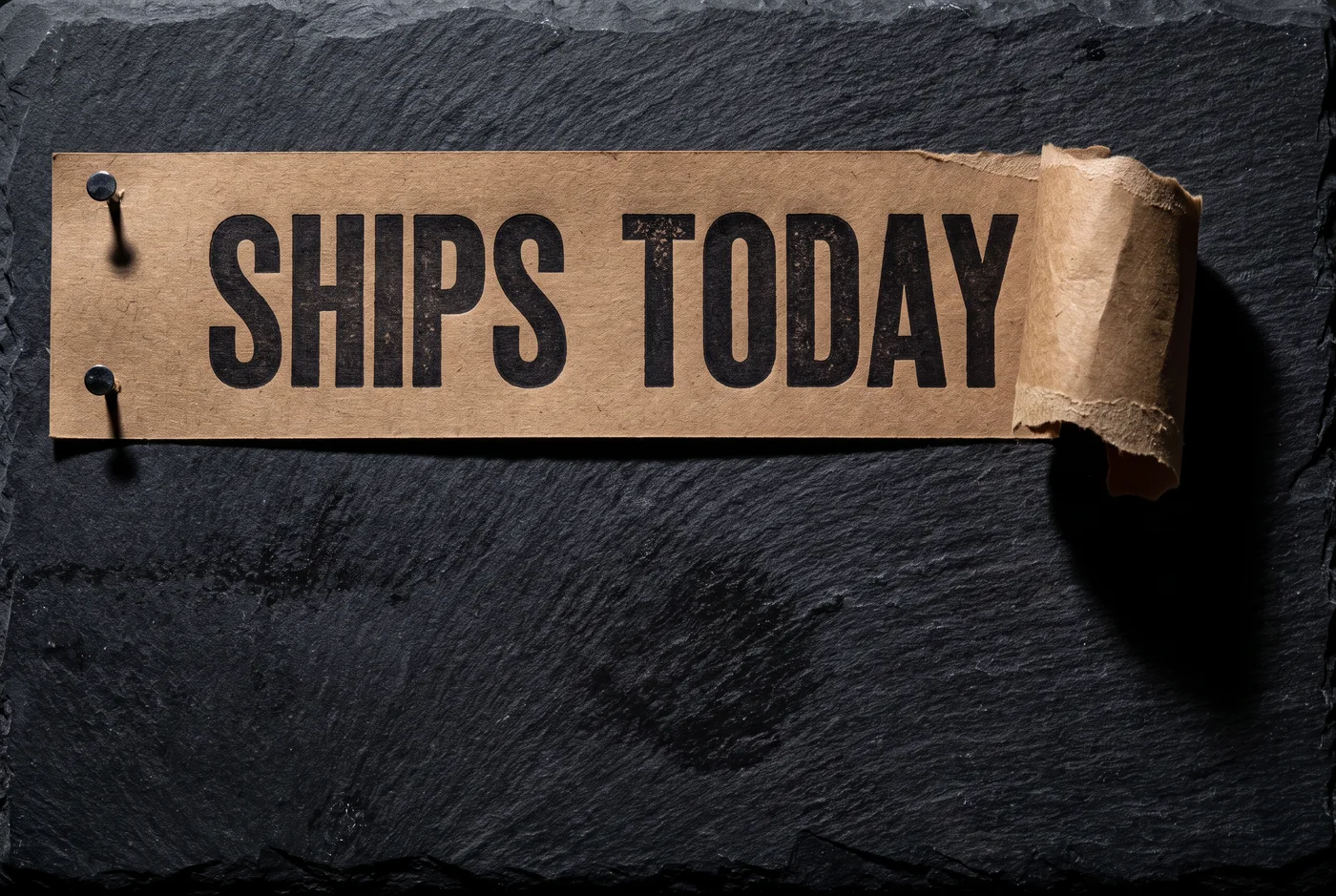 A torn strip of dark kraft paper stretched taut across the top of a black slate slab under hard directional light, letterpressed in two words, one corner ripped away and curling.